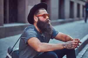 Free photo handsome hipster traveler with a stylish beard and tattoo on his arms dressed in casual clothes with a bag, sits on the sidewalk, resting after a bike ride.