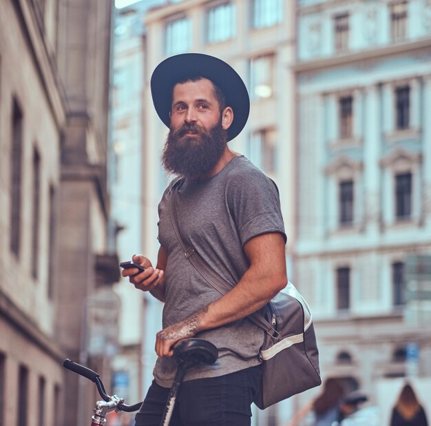 スタイリッシュなあごひげとバッグを持ったカジュアルな服を着た腕にタトゥーを入れたハンサムな流行に敏感な旅行者は、自転車に乗った後、歩道に立って休んでいます。