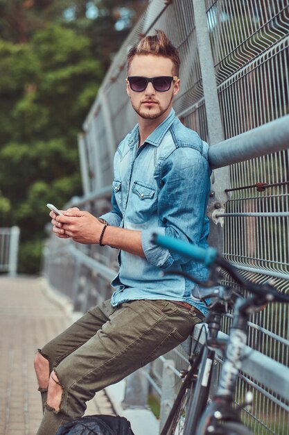 Красивый хипстер со стильной стрижкой в солнцезащитных очках отдыхает после катания на велосипеде, держит смартфон.