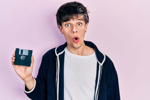 Handsome hipster young man holding floppy disk scared and amazed with open mouth for surprise, disbelief face