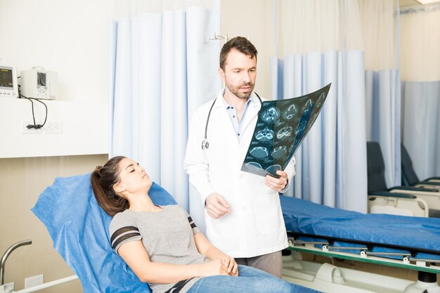 Красивый латиноамериканский врач смотрит на результаты компьютерной томографии пациентки, лежащей на кровати в отделении неотложной помощи
