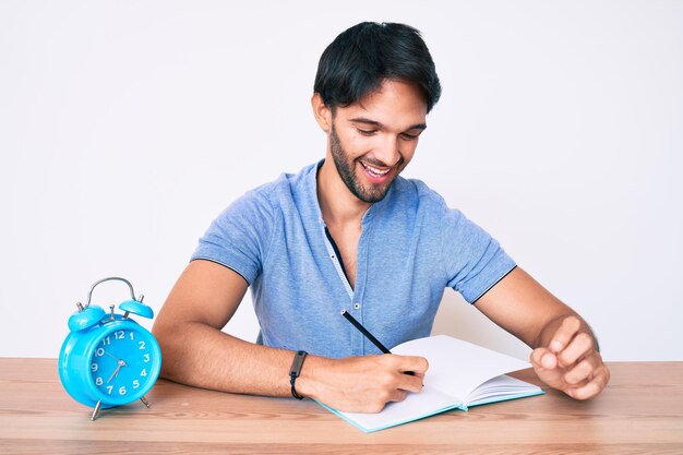 Free photo handsome hispanic man sitting on the table stuying for university looking positive and happy standing and smiling with a confident smile showing teeth