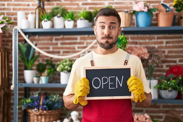 Free photo handsome hispanic man working at florist holding open sign puffing cheeks with funny face mouth inflated with air catching air