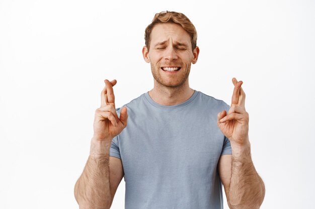 Handsome hopeful ginger guy praying, close eyes and cross fingers for good luck, making wish, waiting for sign, anticipating news, standing over white wall