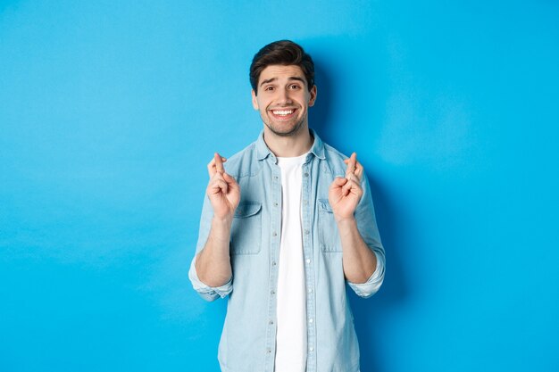 Handsome hopeful man making a wish, crossing fingers and smiling, waiting for results, standing against blue background