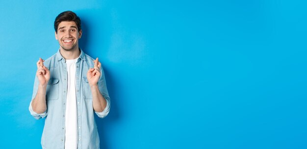 Handsome hopeful man making a wish crossing fingers and smiling waiting for results standing against