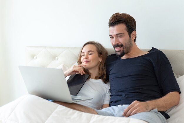잘 생긴 남편과 아름다운 아내는 침대 방에서 컴퓨터 노트북에서 영화를 보는 낭만적 인 부부를 느낍니다.