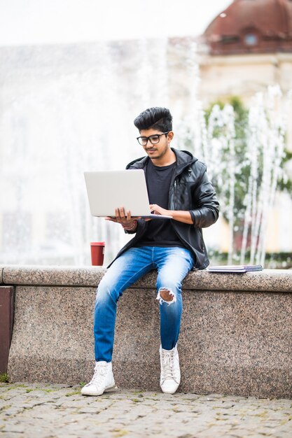 Handsome indian man with laptop while sitting near the fountain in the city center on a day