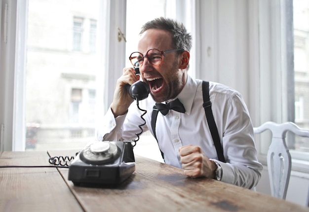 Handsome italian man wearing a white shirt, bowtie sitting near the desk and laughing on the phone