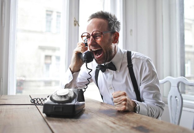 Handsome Italian man wearing a white shirt, bowtie sitting near the desk and laughing on the phone