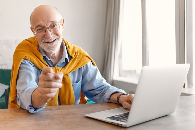 Handsome joyful excited elderly mature man with gray stubble working on laptop in modern office interior sitting at desk by window, smiling and pointing fore finger at camera. Selective focus