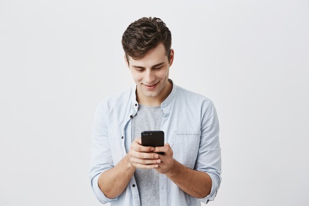Красивый радостный студент с темными волосами, носить синюю рубашку сообщений, набрав сообщение, используя бесплатное приложение onlipe на своем смартфоне, глядя на экран с улыбкой, позирует.
