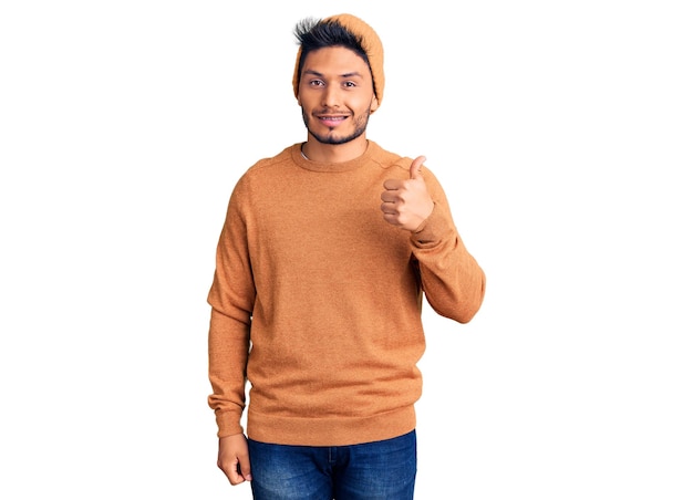 Handsome latin american young man wearing winter sweater and wool hat doing happy thumbs up gesture with hand approving expression looking at the camera showing success