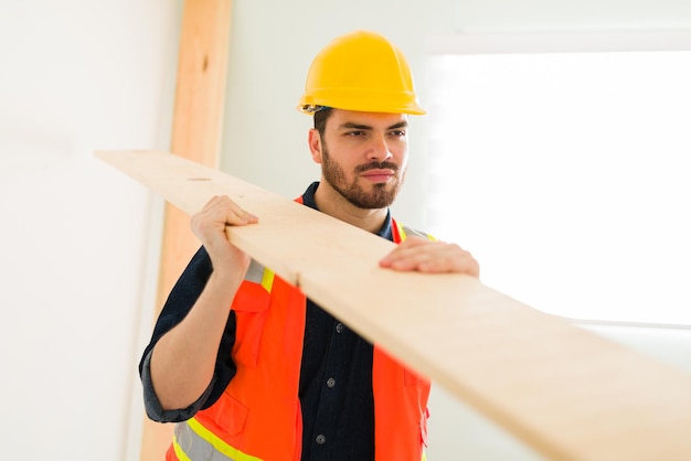 Handsome latin engineer with a helmet and safety vest carrying a wooden panel in the construction site