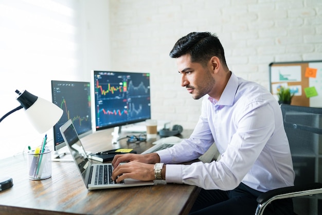 Handsome Latin freelance broker trading through laptop at desk while working from home