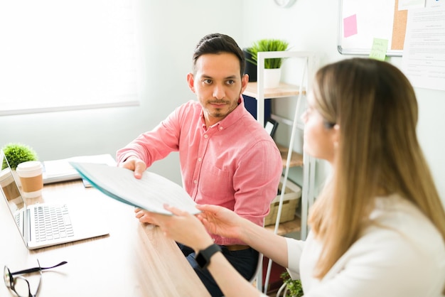 Handsome latin man receiving work papers from his woman business partner. Professional coworkers working on a business report at the office desk