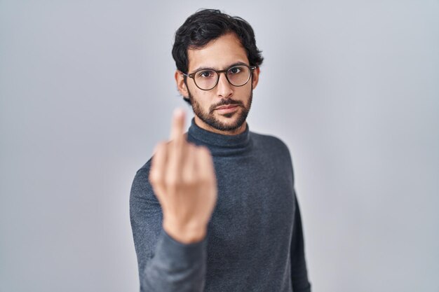 가운데 손가락, 무례하고 무례한 표정을 보여주는 고립 된 배경 위에 서있는 잘 생긴 라틴 남자