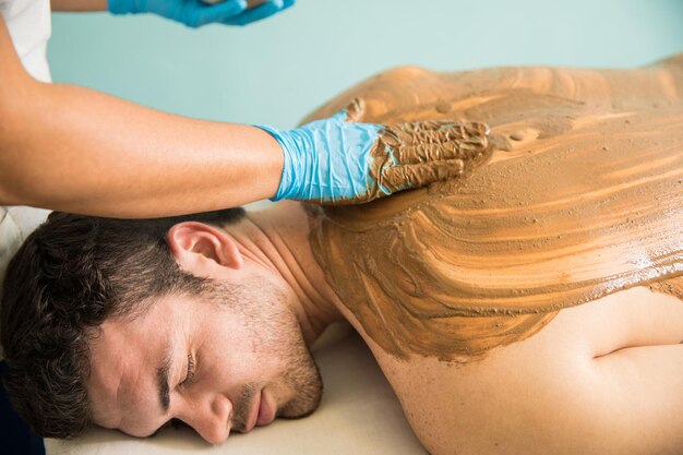 Красивый латинский молодой человек получает массаж и грязевую ванну во время посещения спа-салона
