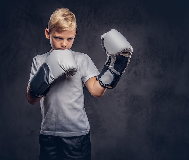 A handsome little boy boxer with blonde hair dressed in a white t-shirt in gloves ready to fight. Isolated on the dark textured background.