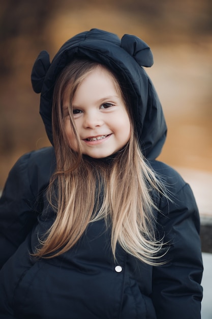 Handsome little girl with long chestnut hair and pretty smile in black jacket goes for a walk in the park in autumn