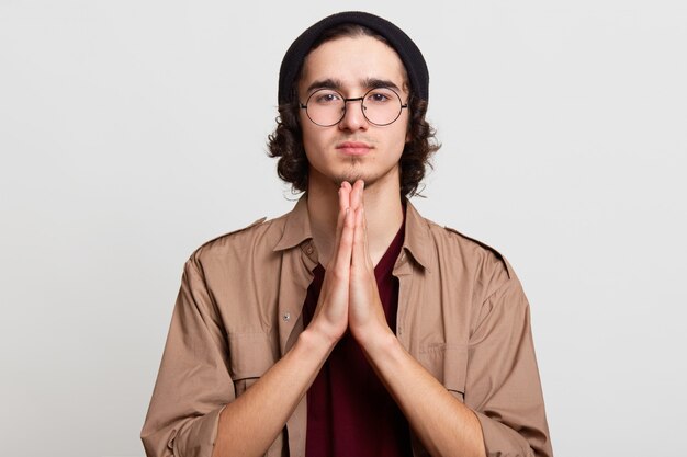 Handsome magnetic young man, putting palms of his hands together, time to prey, pleading, looks peaceful, wearing beige and red shirts, stylish eyeglasses and black hat.