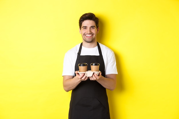 テイクアウトのコーヒーを提供し、笑顔、秩序をもたらし、黄色の背景に黒いエプロンで立っているハンサムな男性のバリスタ。