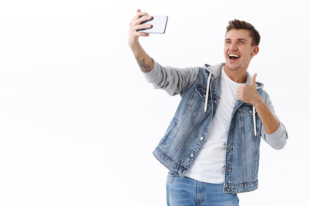 Красивый блогер-мужчина делает селфи на смартфоне, показывает в знак одобрения большой палец вверх, рекомендует хорошее место подписчикам