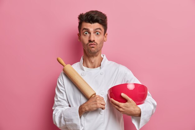 Handsome male chef with surprised expression, cooks food at kitchen, holds rolling pin and bowl, prepares fresh dough, wears white uniform