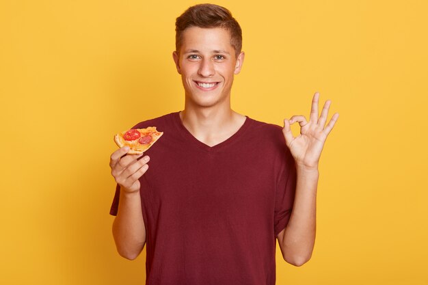 Handsome male dresses casual maroon t shirt holding slice of pizza in hands