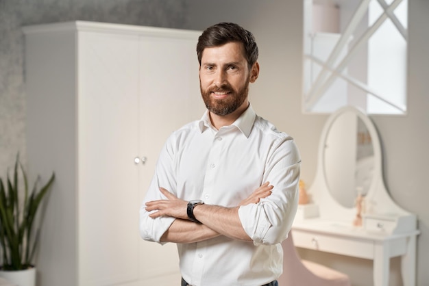 Красивый мужчина-предприниматель в белой рубашке позирует стоя со скрещенными руками в помещении