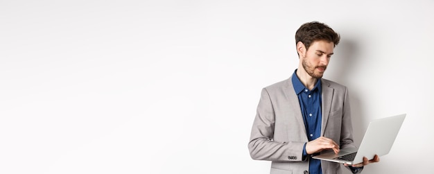 Handsome male entrepreneur working on laptop looking serious at screen standing against white backgr