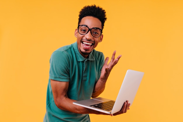 Красивый мужской фрилансер в очках улыбается. Восторженный африканский студент держит ноутбук и выражает счастье.