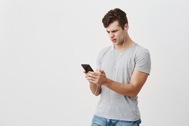Handsome male student holds mobile phone in his hands, looks with confused expression at screen, wears white earphones, frowns face. Caucasian man confused with the message he got from girlfriend.