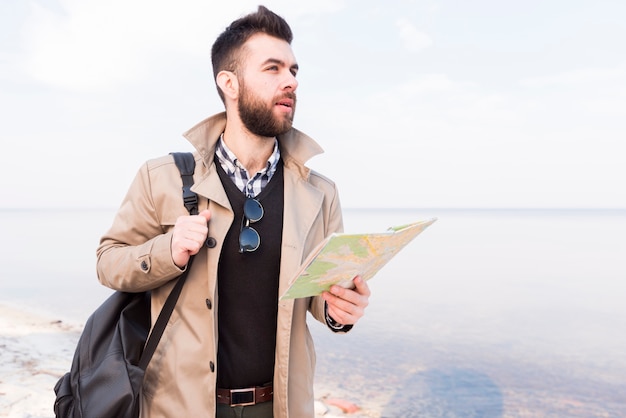 Красивый мужчина путешественник, стоя возле моря, держа карту в руке, глядя