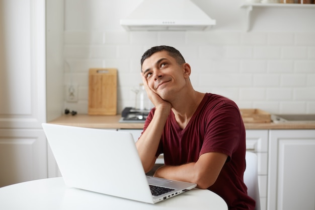 Handsome male wearing burgundy casual style t shirt sitting in front of portable computer at table in kitchen, dreaming about vacation, imagines how will be spend weekend.