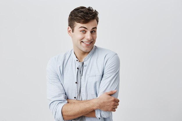 Handsome male with dark hair looking  with friendly smile, enjoying good day and leisure time indoors, keeping arms folded. Young european man feeling happy and carefree while relaxing.