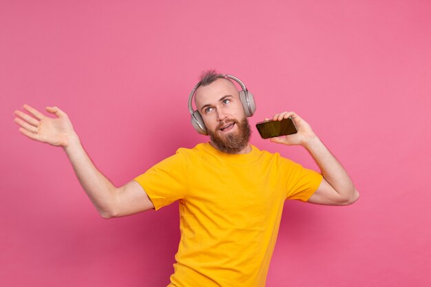 Handsome man in casual dancing with mobile phone and headphones isolated on pink background