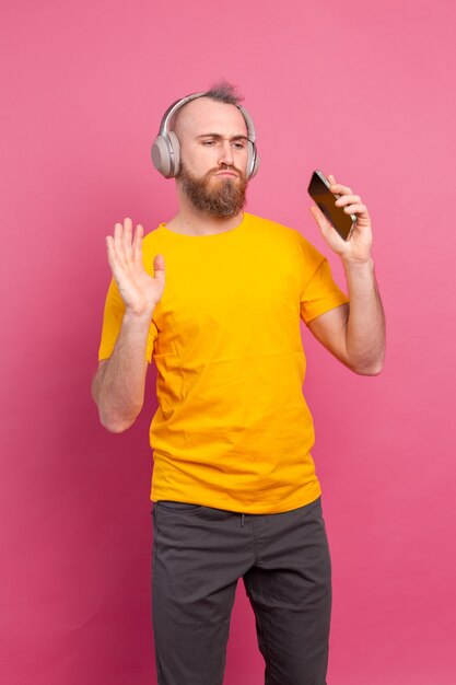 Handsome man in casual dancing with mobile phone and headphones isolated on pink background