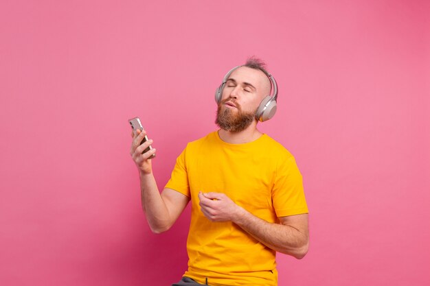 Handsome man in casual dancing with mobile phone and headphones isolated on pink background