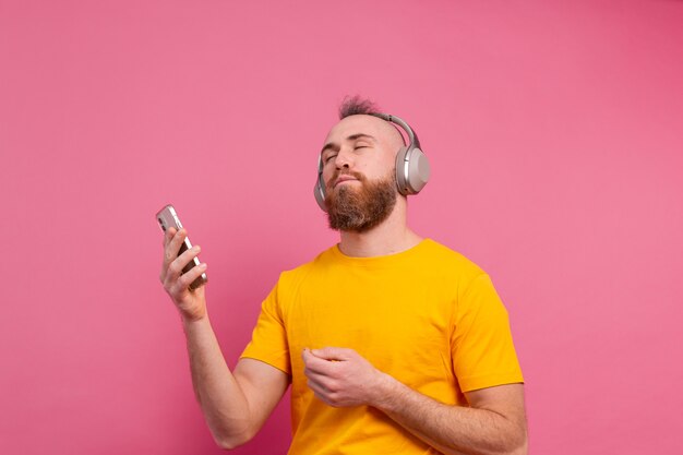 Handsome man in casual dancing with mobile phone and headphones isolated on pink background