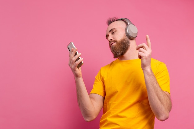 Handsome man in casual dancing with mobile phone and headphones isolated on pink background