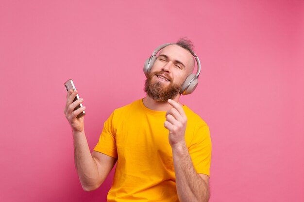 Handsome man in casual dancing with mobile phone and headphones isolated on pink background