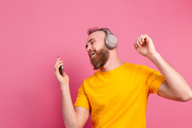 Handsome man in casual dancing with mobile phone and headphones isolated on pink background