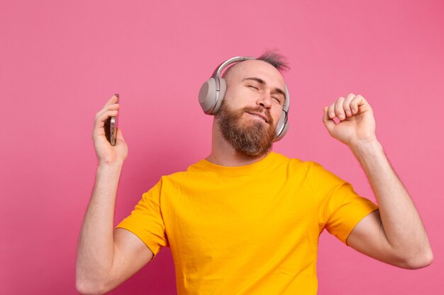 Handsome man in casual dancing with mobile phone and headphones isolated on pink background