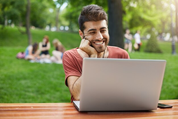 Красивый мужчина соединяет Wi-Fi в парке и друга по видеосвязи с ноутбуком
