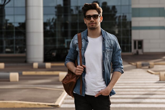 Handsome man in denim jacket and white tshirt poses outside Portrait of brunette guy in sunglasses holding bag