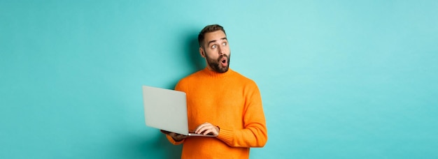 Бесплатное фото Красивый мужчина, делающий покупки в интернете, задумчиво смотрит вверх, используя ноутбук, стоящий над голубым