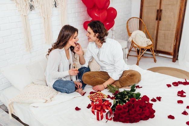 Handsome man feeding girlfriend with chocolate candy and drinking champagne while sitting on the bed and home, st valentines day concept