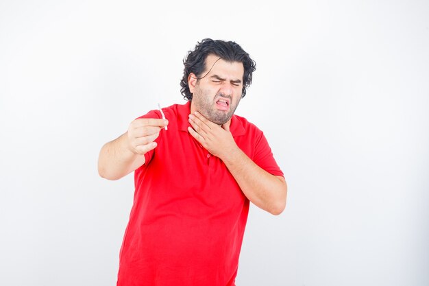 Handsome man holding cigarette, holding hand on neck, coughing in red t-shirt and looking displeased , front view.