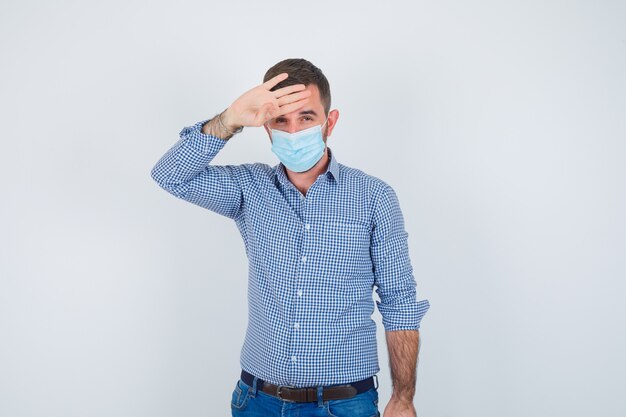 Handsome man holding hand on head, checking his temperature in shirt, jeans, mask and looking exhausted. front view.